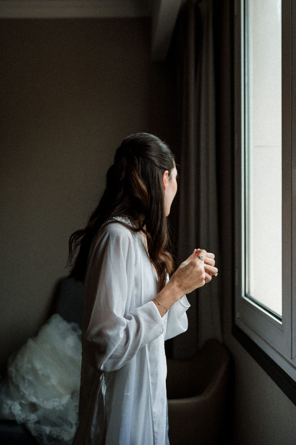 A person in a white dress stands by a window in a dark room creating a moody, contemplative atmosphere.