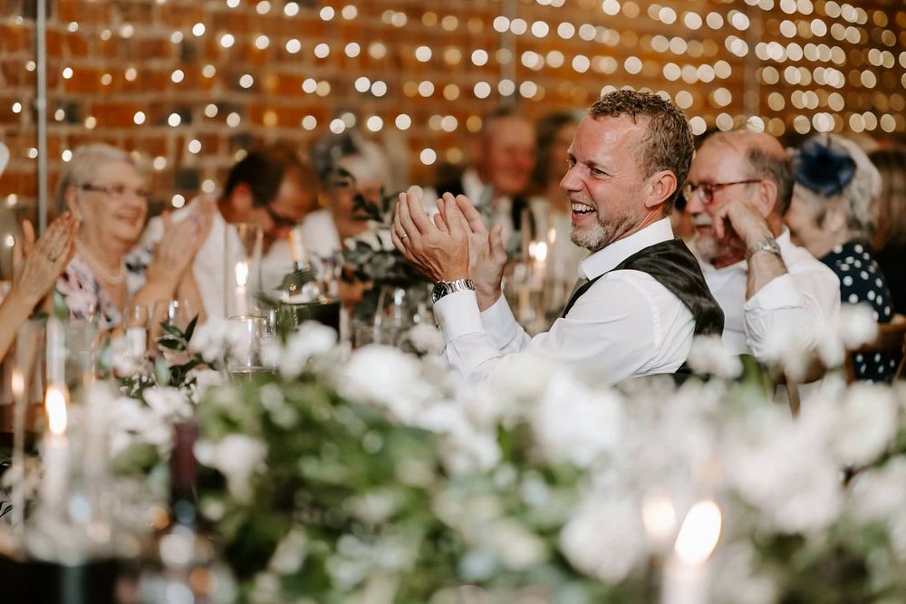 Wedding reception guests applaud during celebration with string lights and white floral decorations in background.