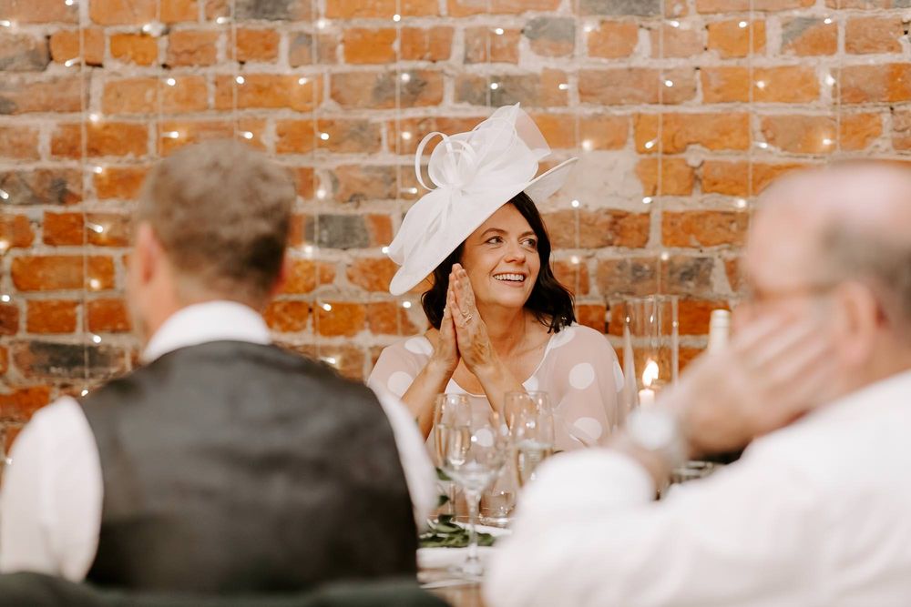 Wedding guests seated at long tables in a rustic brick-walled venue share conversation and laughter during reception.