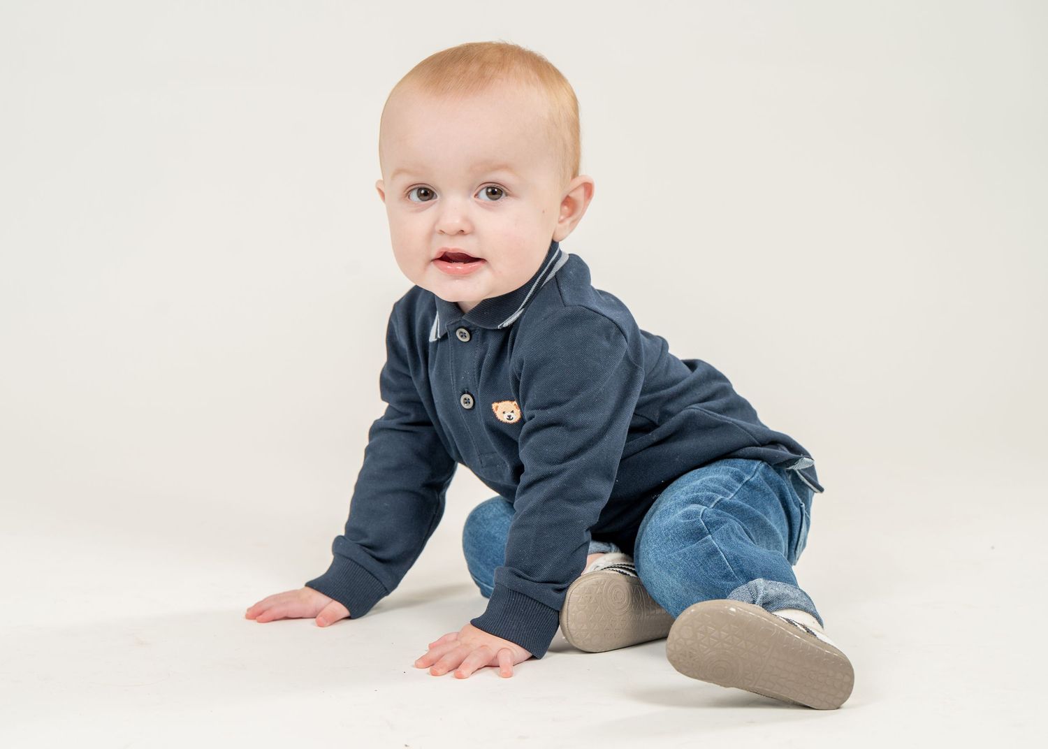 Baby in a navy blue sweater and jeans crawls and plays on a white studio backdrop.