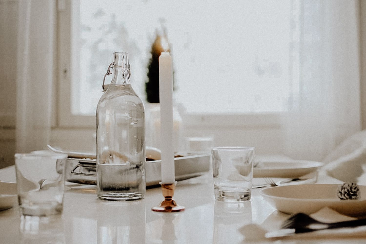 A minimalist table setting with a glass bottle, lit candle, and water glasses in soft natural light.