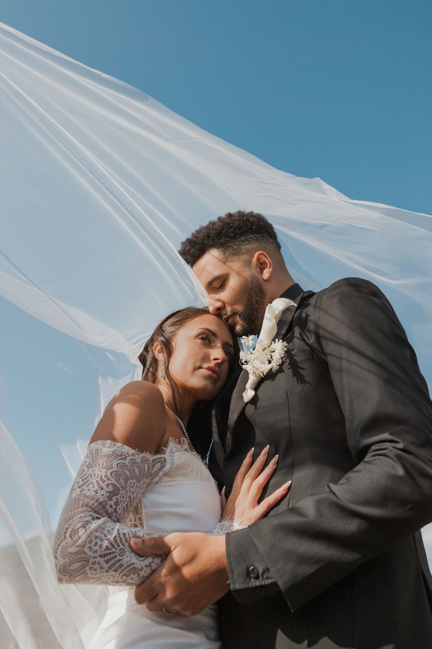 A romantic wedding photo shows a couple embracing under a flowing white veil against a bright blue sky.