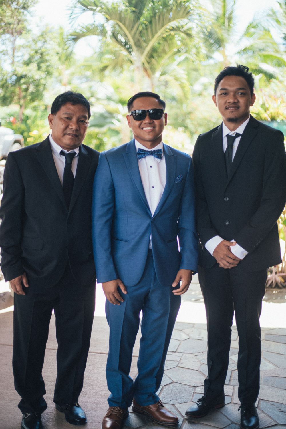 Wedding party in black and blue suits posing together against a palm tree backdrop.