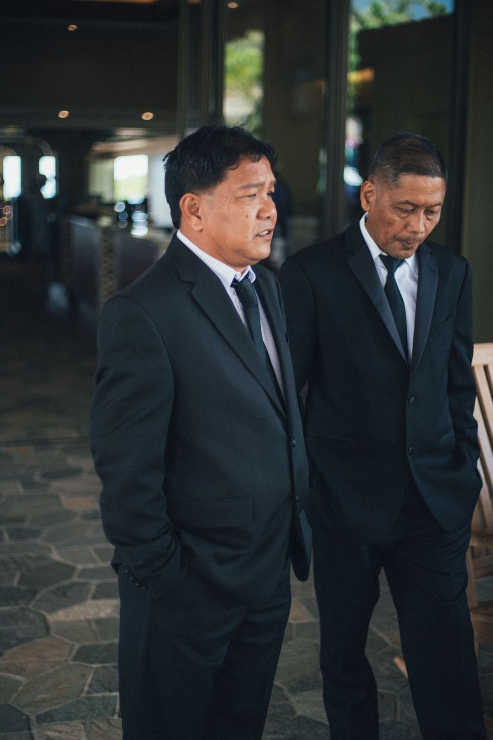 Two men in black suits and ties stand together in a formal setting with stone flooring.