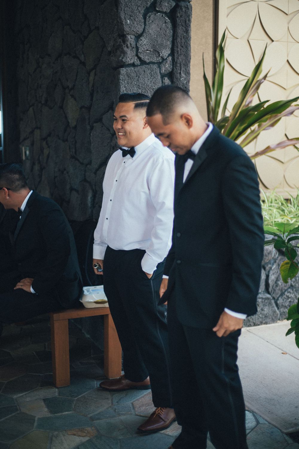 Two men in formal attire stand near a stone wall with tropical plants in the background.