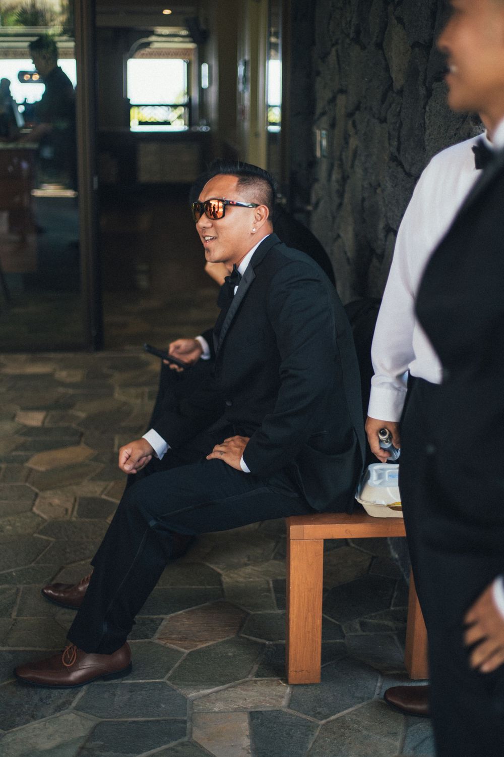 Man in formal attire sits on wooden bench while laughing at outdoor venue.