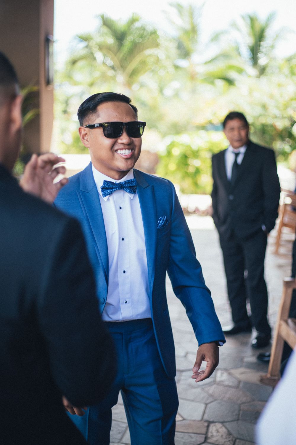 Man in bright blue suit and bow tie smiles while wearing sunglasses at outdoor event.