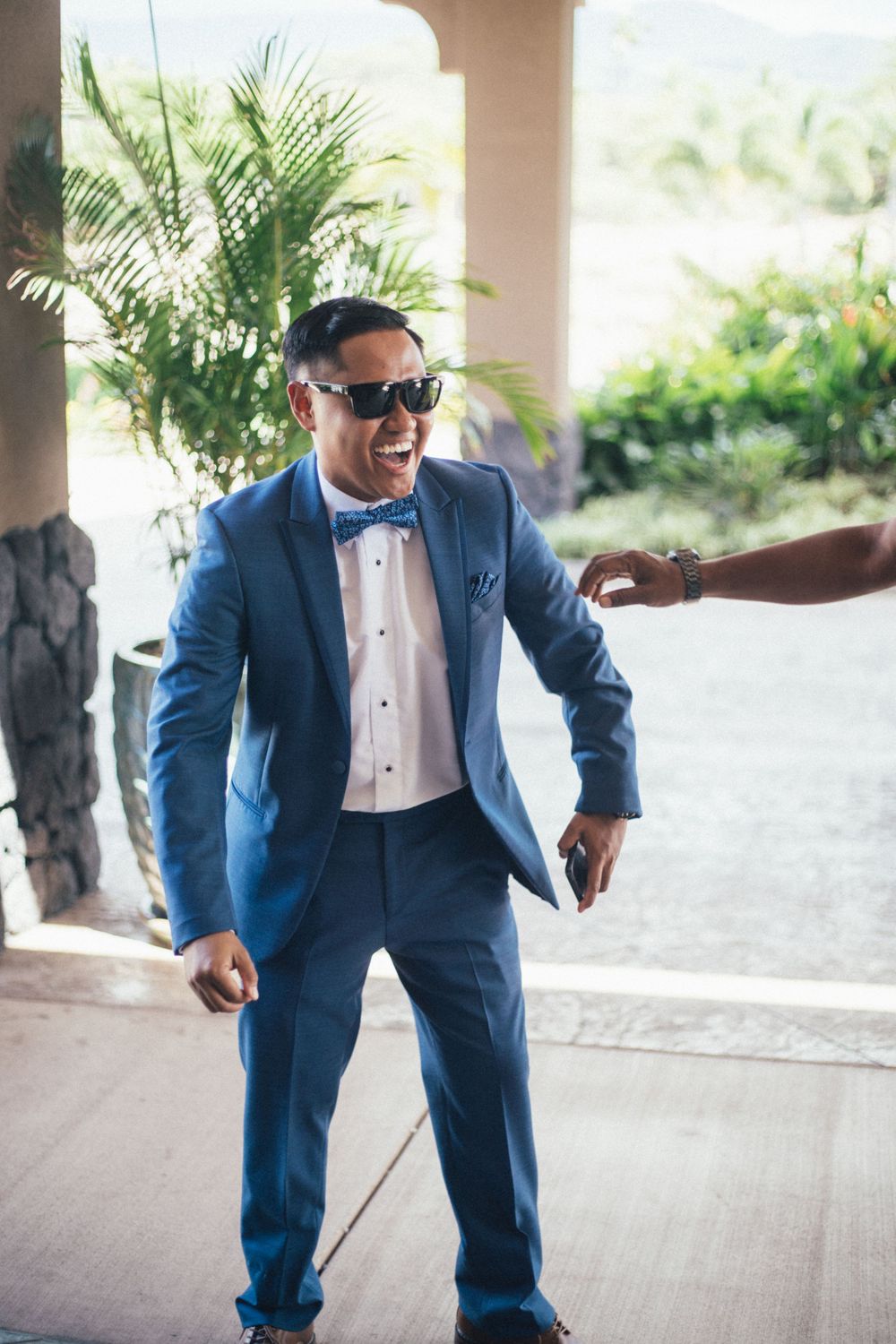 Man in blue tuxedo and bow tie dances joyfully while wearing sunglasses at an outdoor venue.