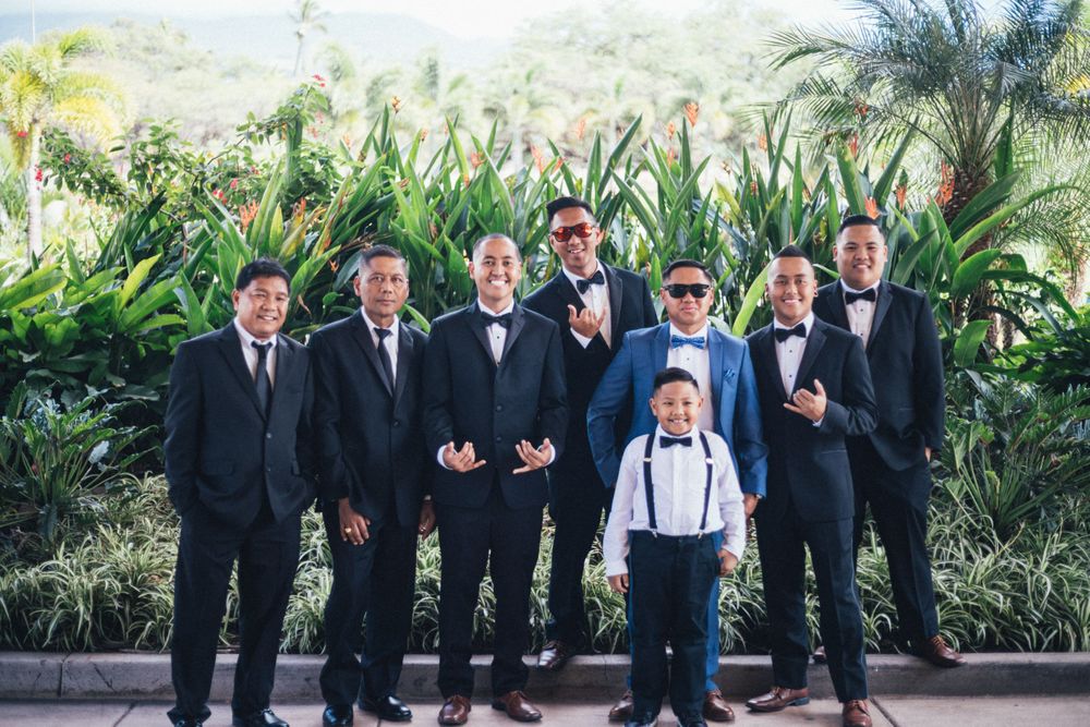 Groomsmen in formal suits and bowties posing together outdoors with tropical landscaping.