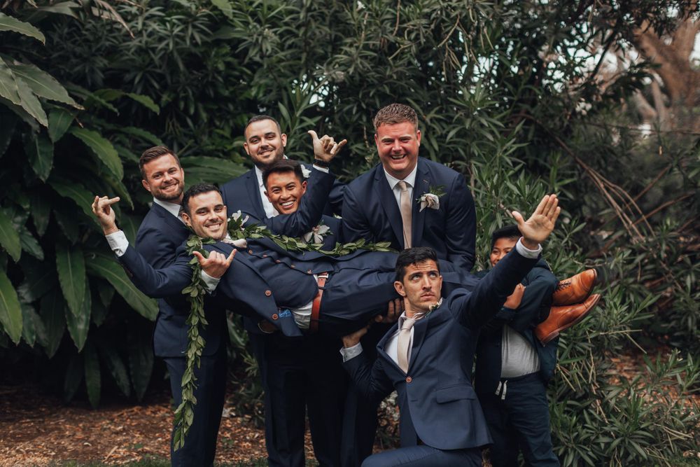 Groomsmen in dark suits pose together outdoors during wedding celebrations.