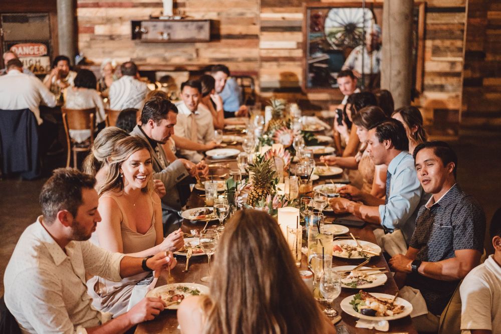 Wedding reception dinner with guests seated at long tables in rustic wooden venue.