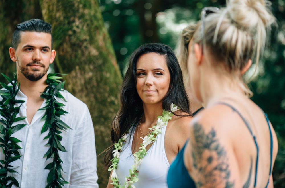 Three people wearing traditional Hawaiian leis stand together in a tropical forest setting.