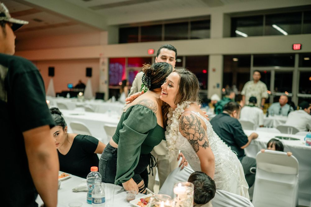 Wedding guests share an emotional embrace at a reception with white chairs and tables in the background.