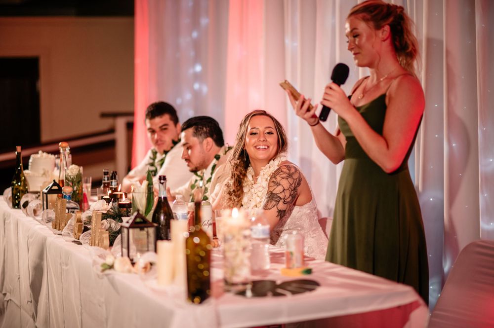 Wedding party at head table listens to speech during reception with pink lighting and white decor.