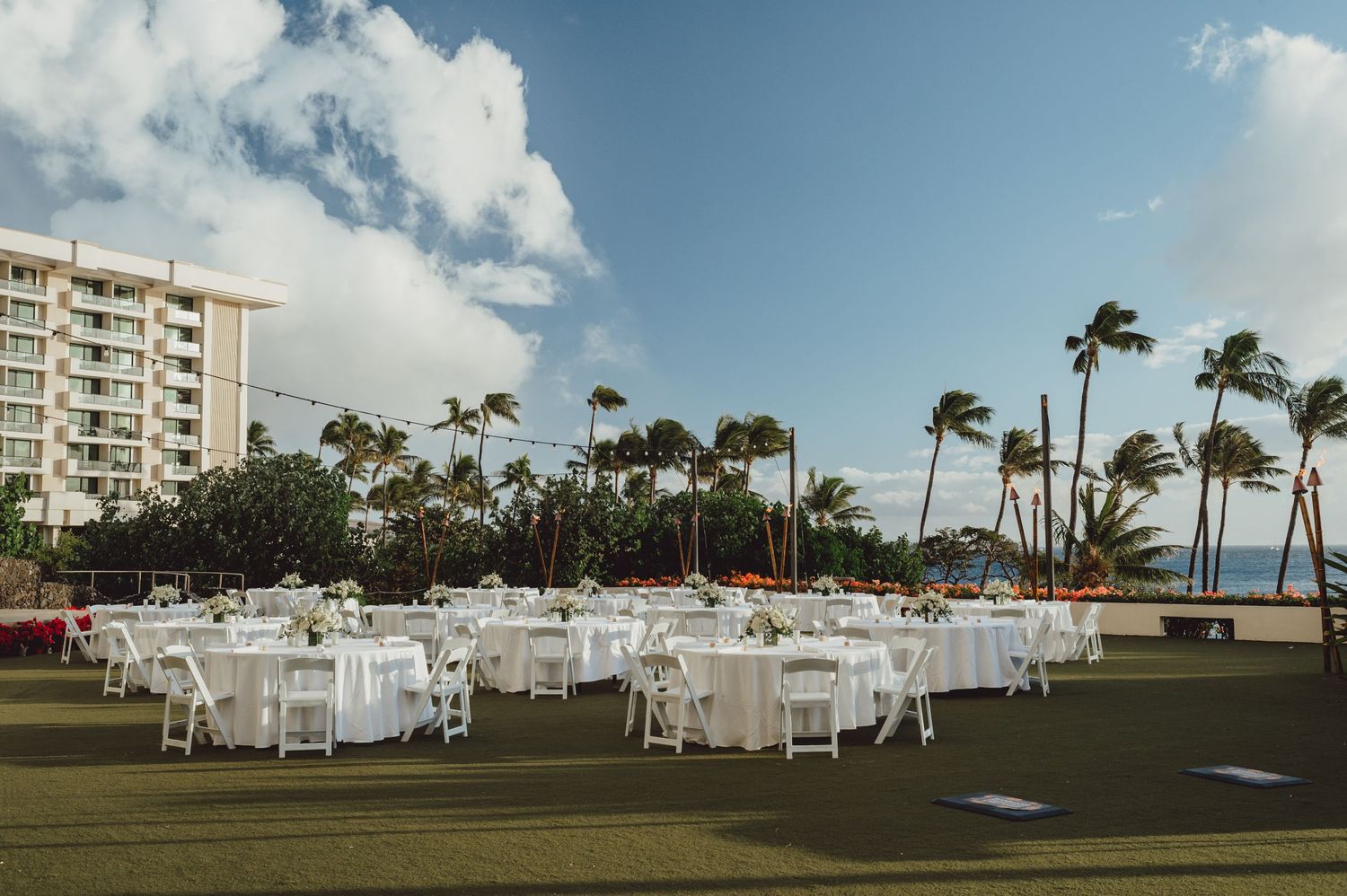 White tables and chairs set up for an outdoor wedding reception on a lawn with palm trees and high-rise buildings in Hawaii.