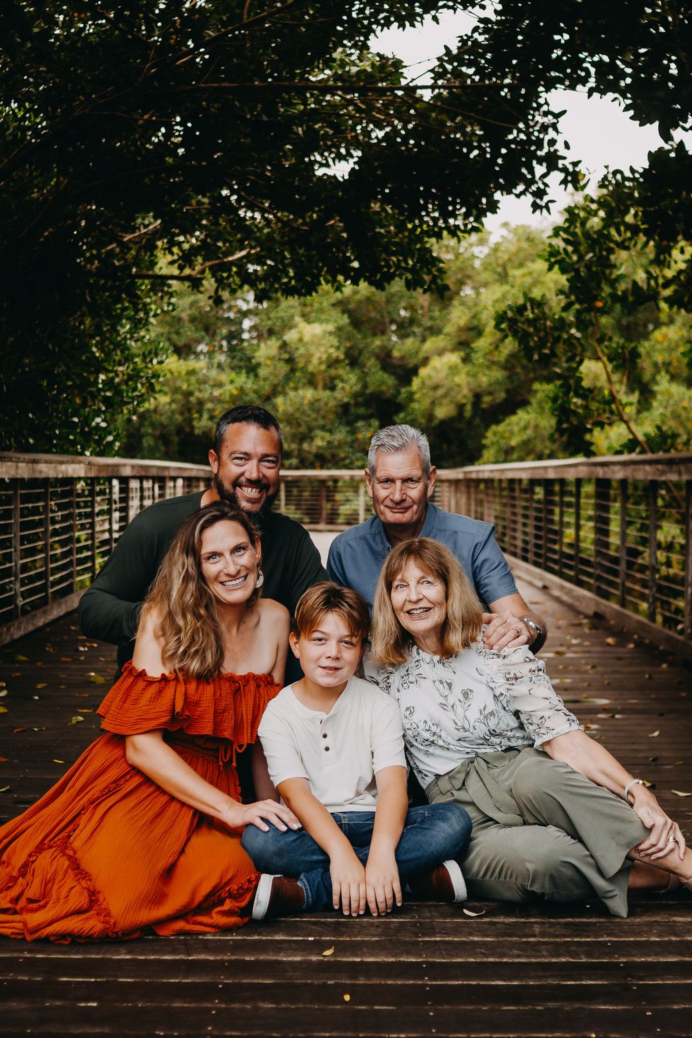 A family poses together on a wooden bridge surrounded by lush green trees on a sunny autumn day.