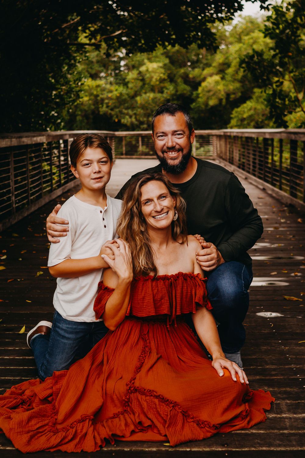 A family poses together on a wooden bridge walkway, with the woman wearing a flowing red dress against a natural backdrop.