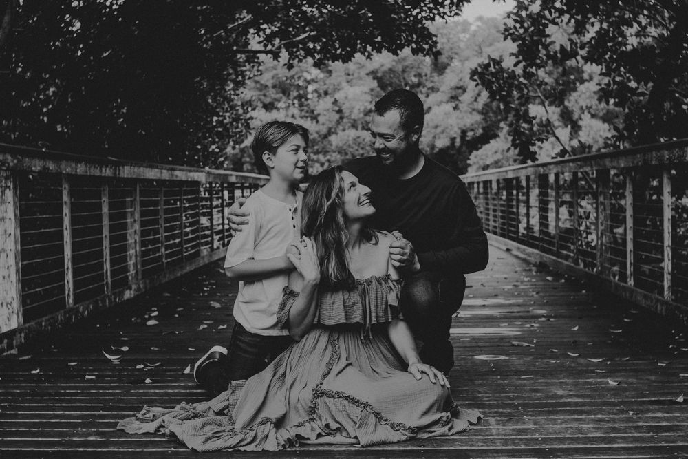 A couple sits together on a wooden bridge surrounded by trees in a romantic black and white photograph.