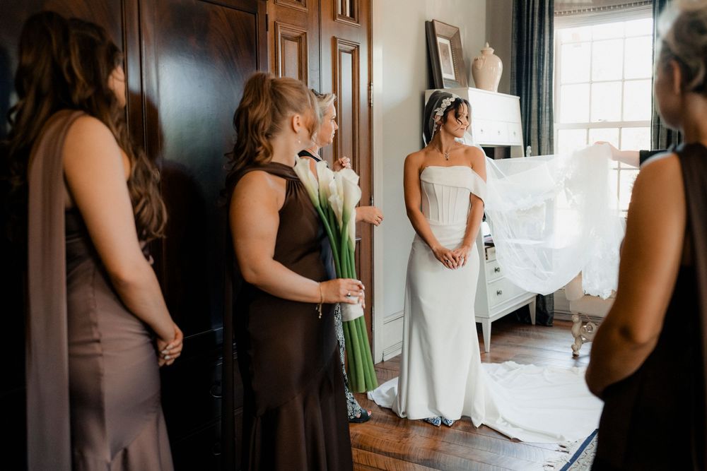 Bride in white corset gown with veil surrounded by bridesmaids in dark dresses indoors.