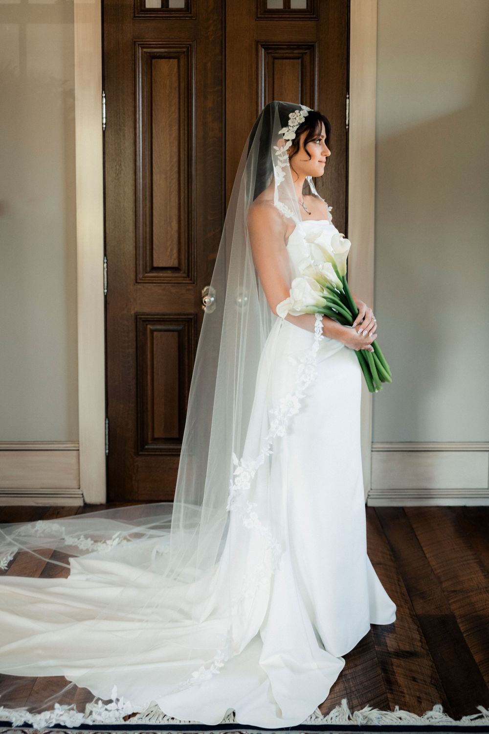 Bride in white strapless gown with long lace-trimmed cathedral veil holding calla lily bouquet by wooden doors.