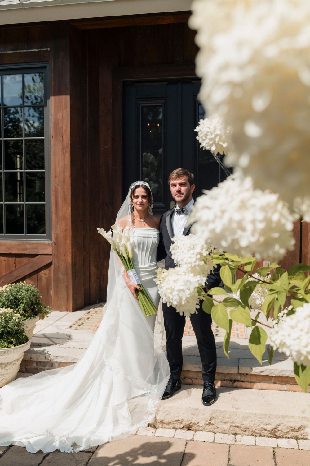 Bride and groom posing outdoors by rustic wooden building surrounded by white hydrangeas and calla lily bouquet.