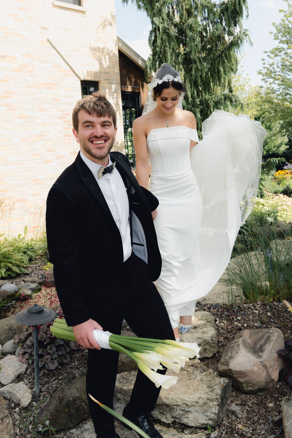 Smiling groom in black tuxedo holds calla lilies while bride in strapless gown stands on garden rocks behind him.
