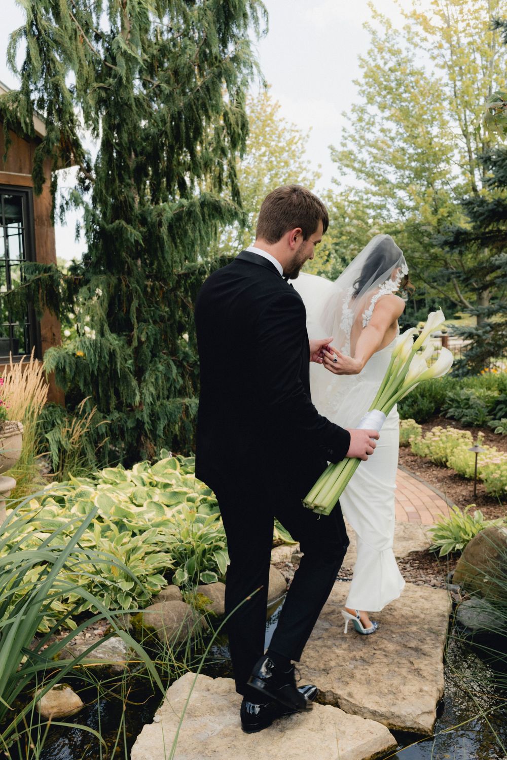 Groom in black suit walks with bride in white gown and veil through lush garden landscape holding calla lilies.