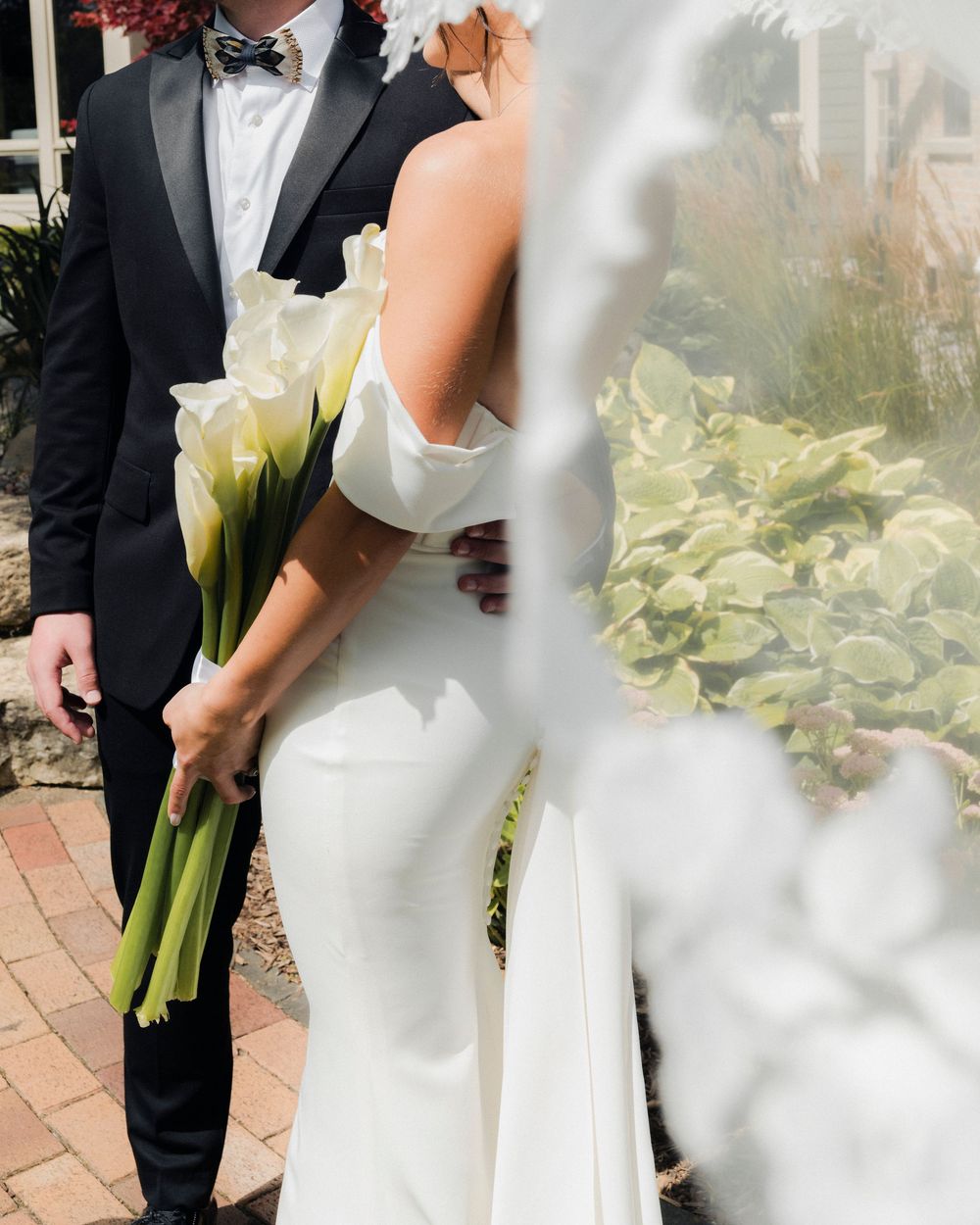 Close-up of bride and groom embracing outdoors, groom holding white calla lily bouquet with veil flowing in foreground.