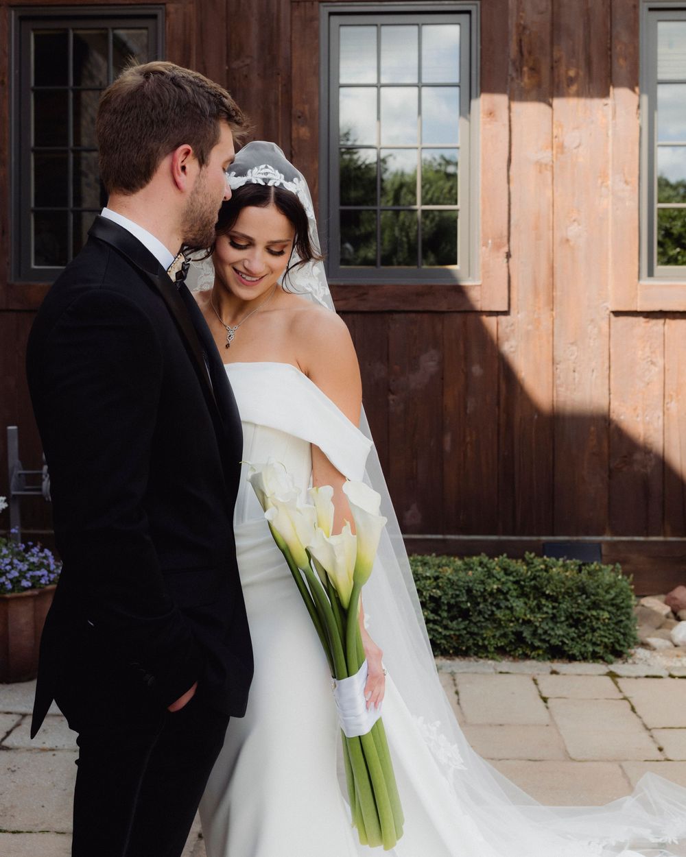 Bride in strapless white gown with lace veil leans into groom in black tuxedo outside rustic wooden barn venue.