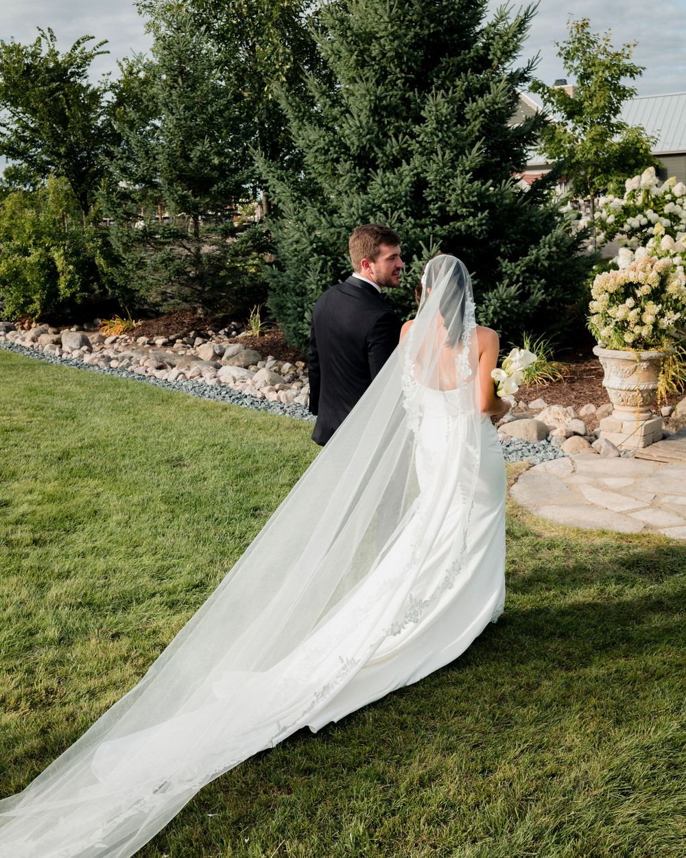Bride and groom walking across green lawn, bride's long cathedral veil trailing behind them near garden landscaping.