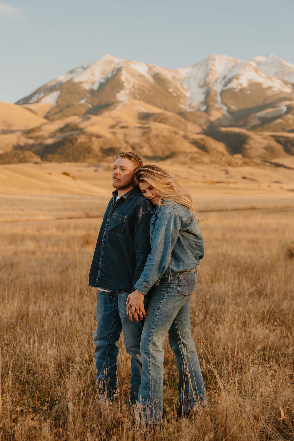 A couple embrace in a golden field with snow-capped mountains in the background at sunset overlooking mountains in Chico, Montana.