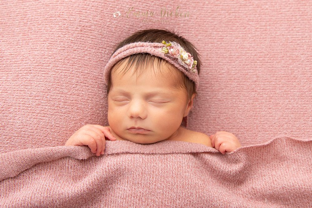 Newborn baby girl poses with flower headband tucked in bed