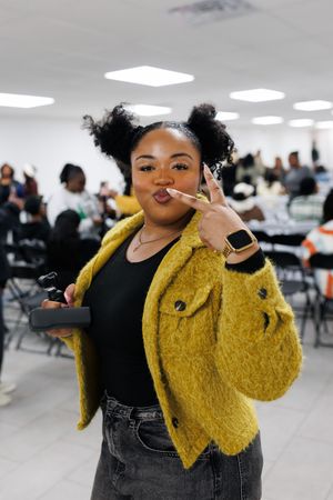 A person in a yellow fuzzy jacket and black overalls poses playfully in a cafeteria setting.