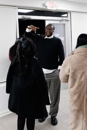 A person in a black sweater and glasses stands near an exit door while others gather in a hallway.