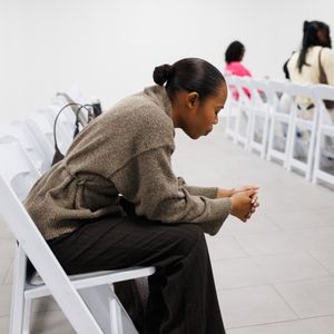 People in formal attire seated in white folding chairs at an indoor event.