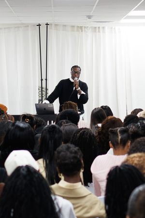 Speaker in dark suit addresses a packed audience in a bright white room.