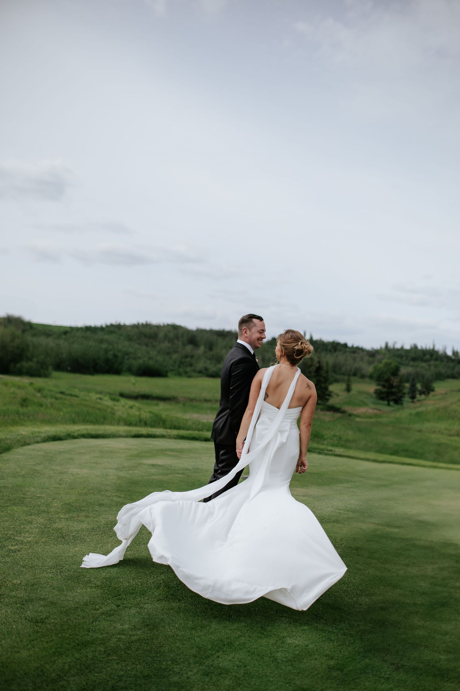 A couple in wedding attire dances and twirls across a scenic green golf course under cloudy skies.