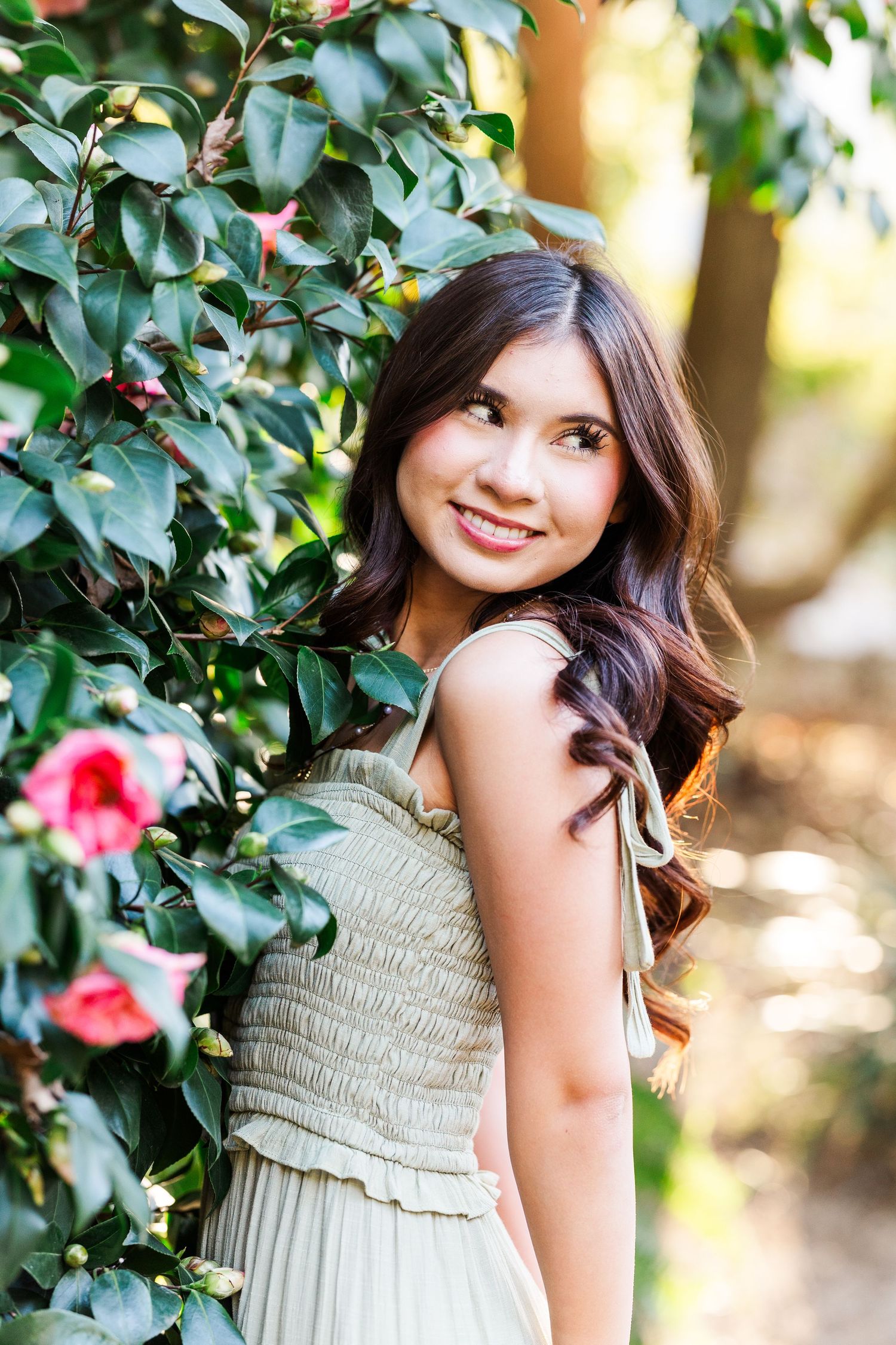 Portrait of someone in a beige smocked dress standing among flowering camellia bushes in a garden setting.
