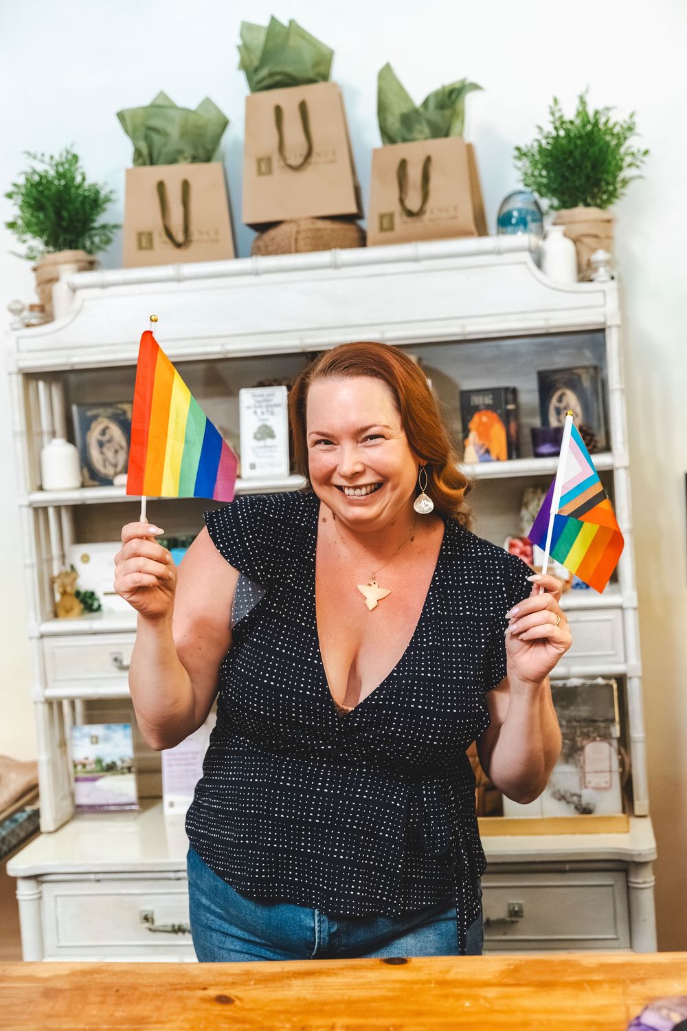 Someone holds rainbow pride flags while smiling in front of a white shelving unit decorated with shopping bags and plants.