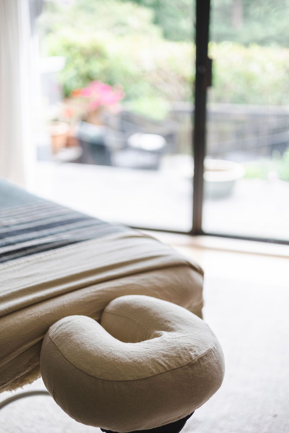 A beige neck support pillow rests on a massage table near a window with soft natural lighting.