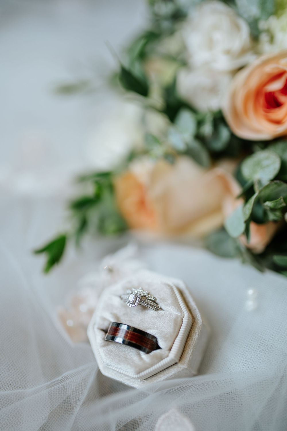 Wedding rings nestled in delicate white tulle fabric with soft peach roses and greenery in the background.