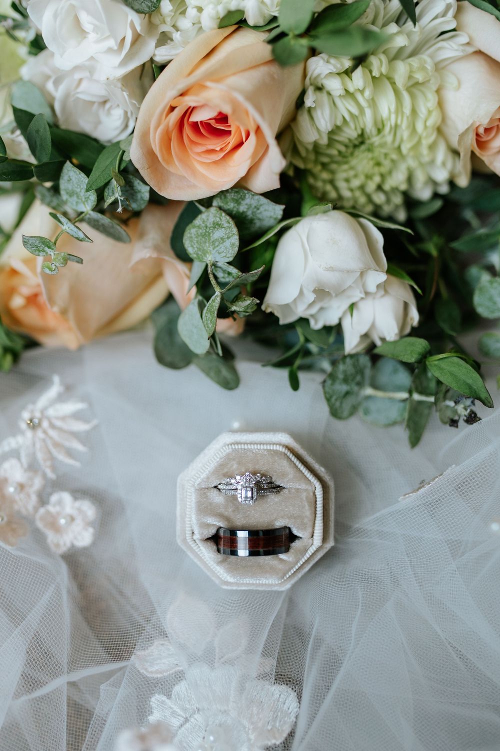 Wedding rings displayed on white tulle fabric with peach roses and greenery in the background.