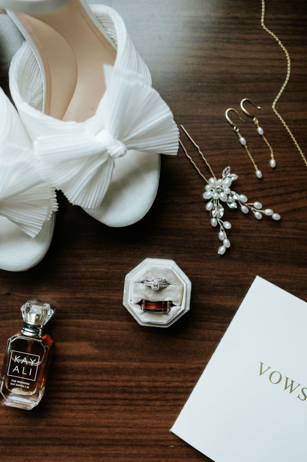 Wedding details including perfume bottles, jewelry, and vows book arranged on dark wooden surface.