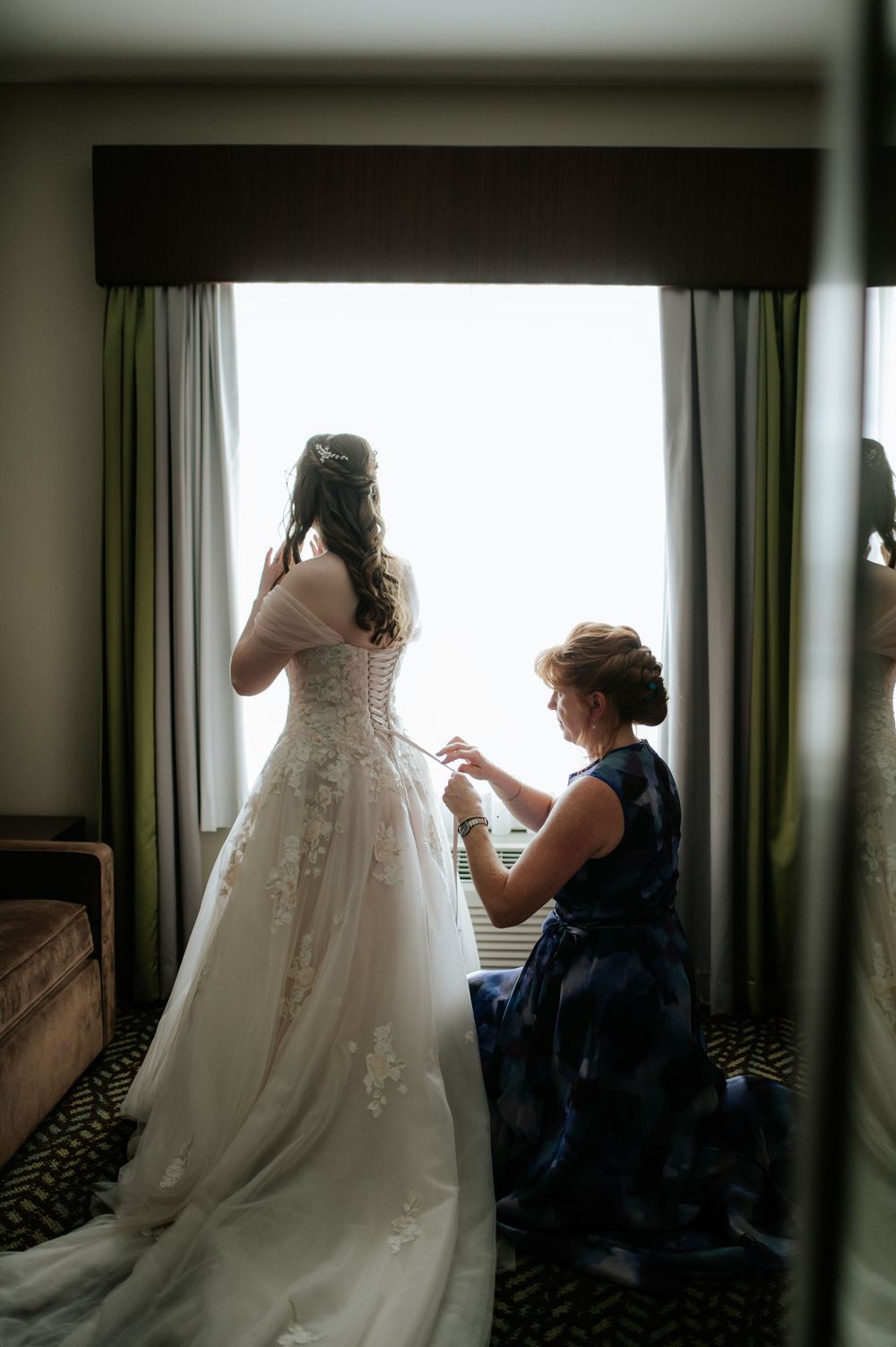 A wedding dress being buttoned up by someone in a navy dress near a window with green curtains.