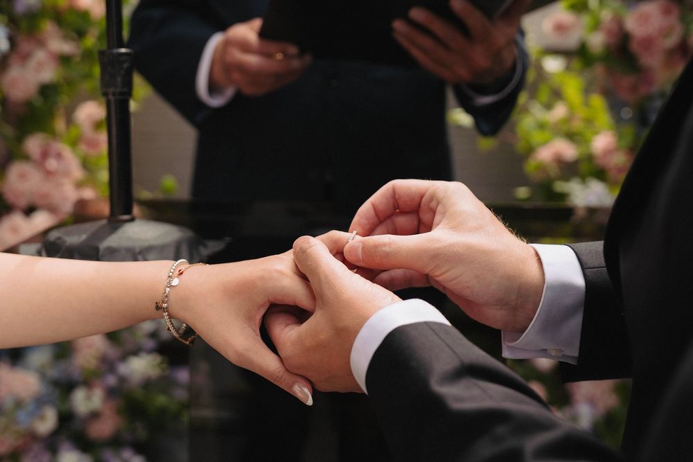 Close-up of wedding ceremony hands exchanging rings with pink floral decorations in background.