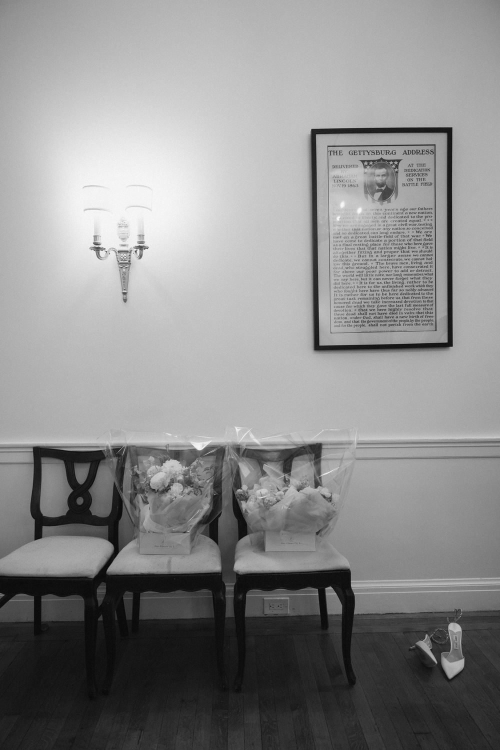 Black and white interior shot of elegant chairs against a wall with framed artwork and sconces.