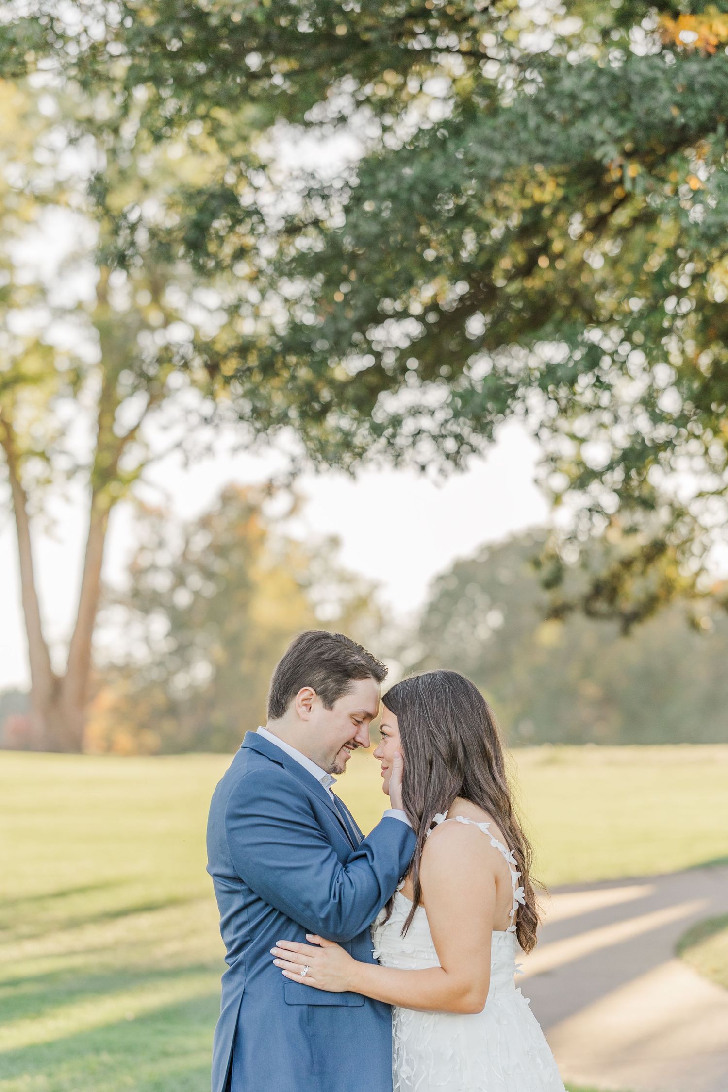 A couple shares an intimate moment outdoors on a sunny day with trees and greenery in the background.