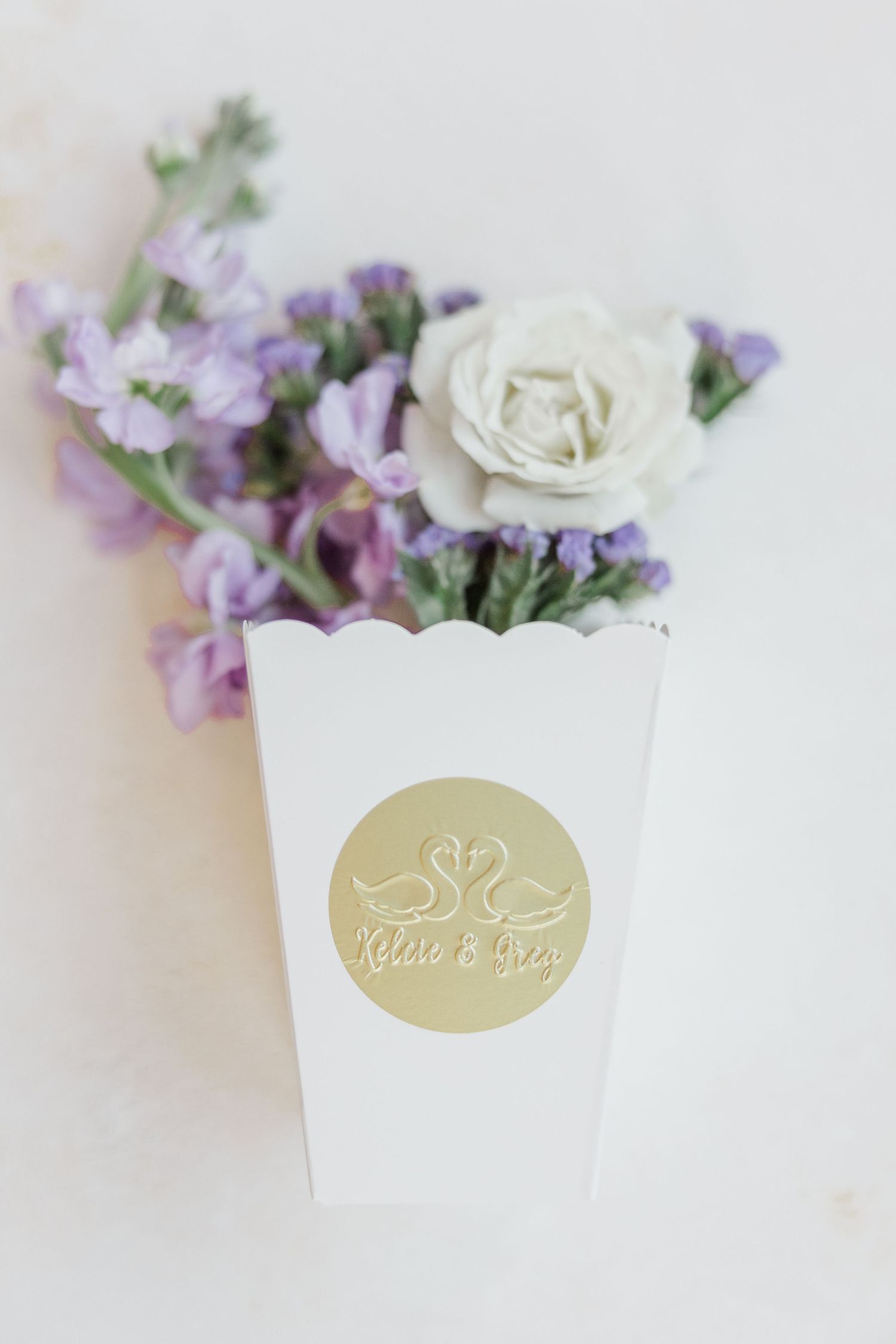 Elegant wedding invitation details with gold foil stickers and delicate purple and white flowers arranged on white background.