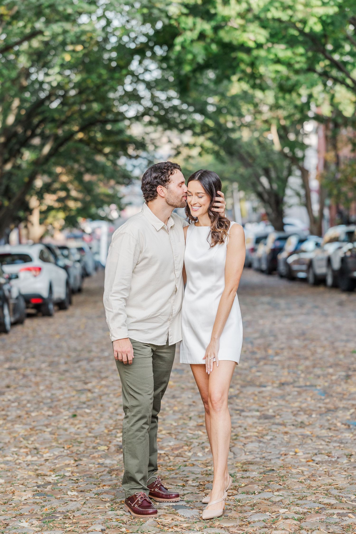 A romantic couple embraces on a cobblestone street lined with parked cars and autumn leaves.