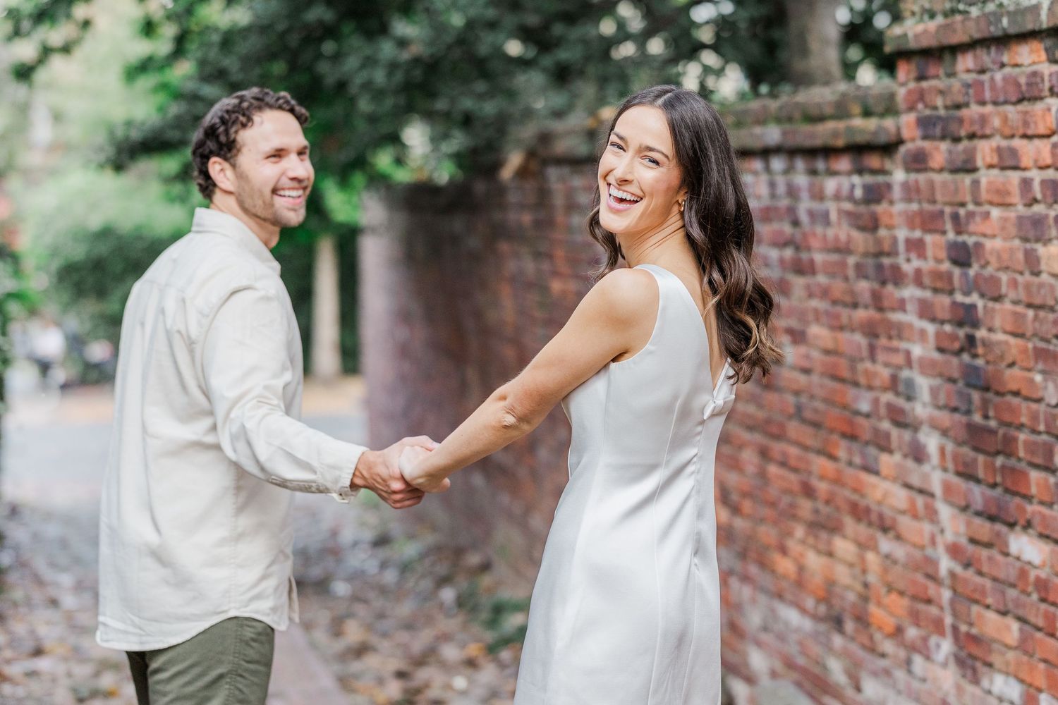 A couple in white wedding attire dances and laughs together along a rustic brick wall in a garden setting.
